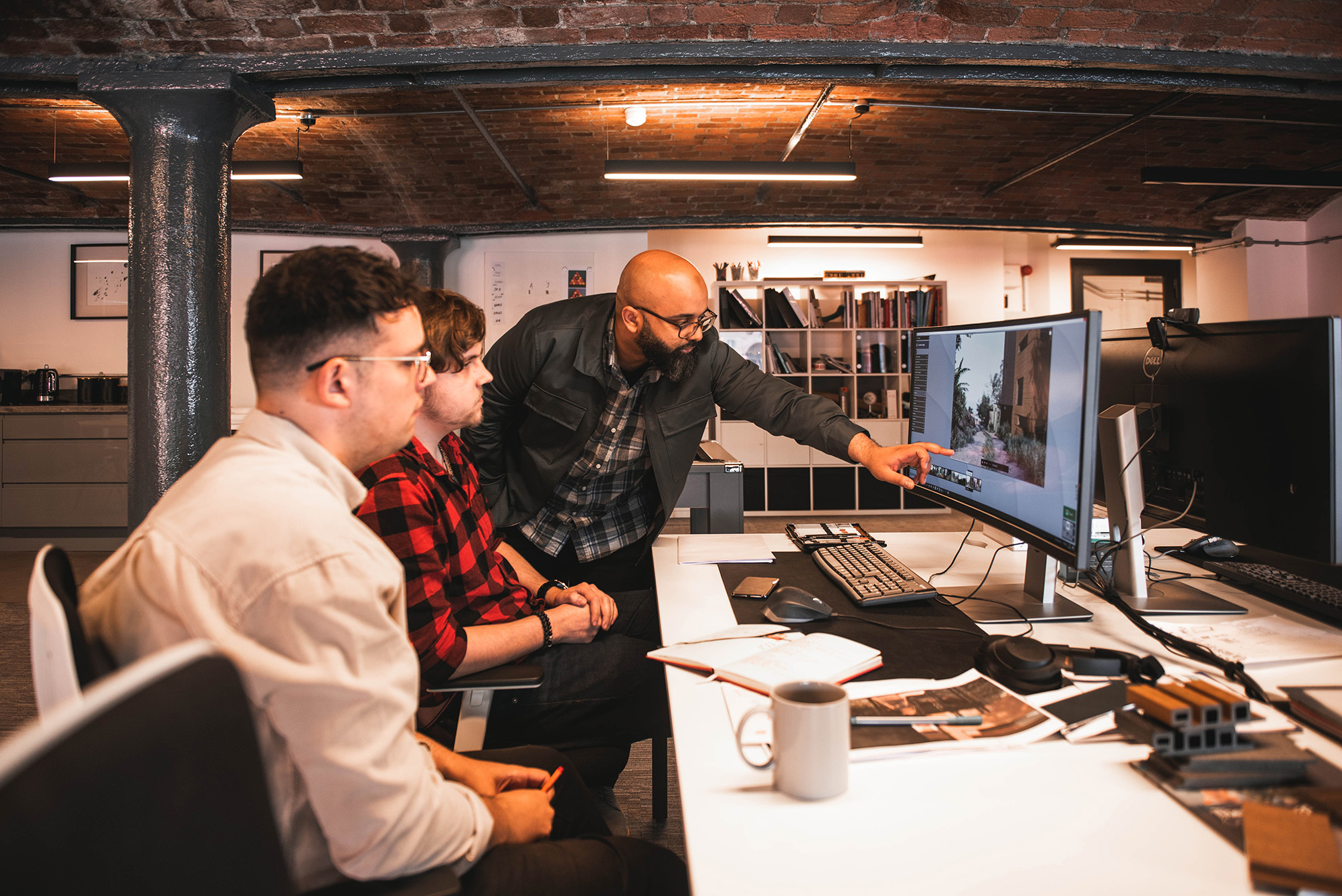 Architect mentor guiding two colleagues through a visualization project on a curved monitor in a brick-walled design studio